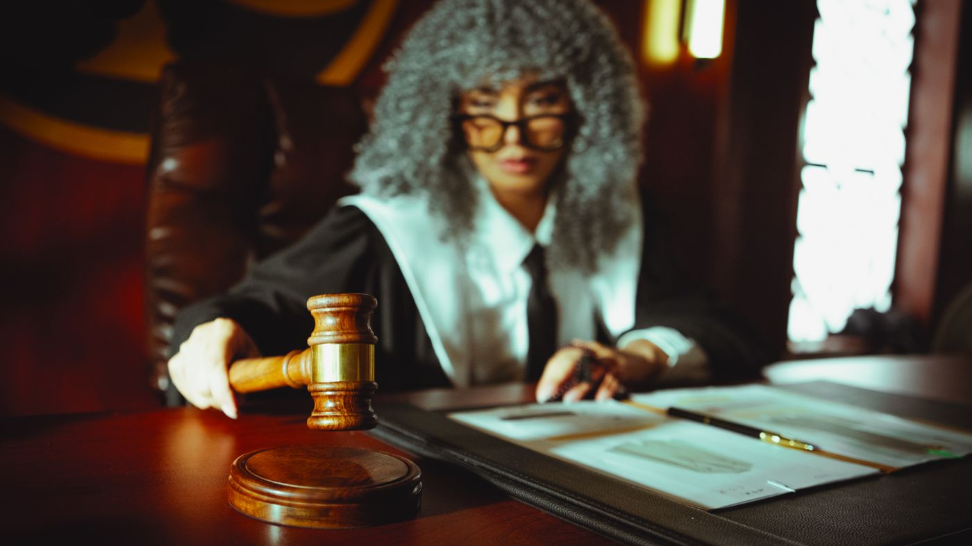 A judge in black robes and glasses sits at a courtroom bench, resting one hand on a wooden gavel while reviewing an open folder of legal documents.