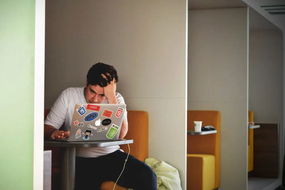 A man sits at a small cafe table, holding his head in one hand with a frustrated expression as he stares at a sticker-covered laptop screen.