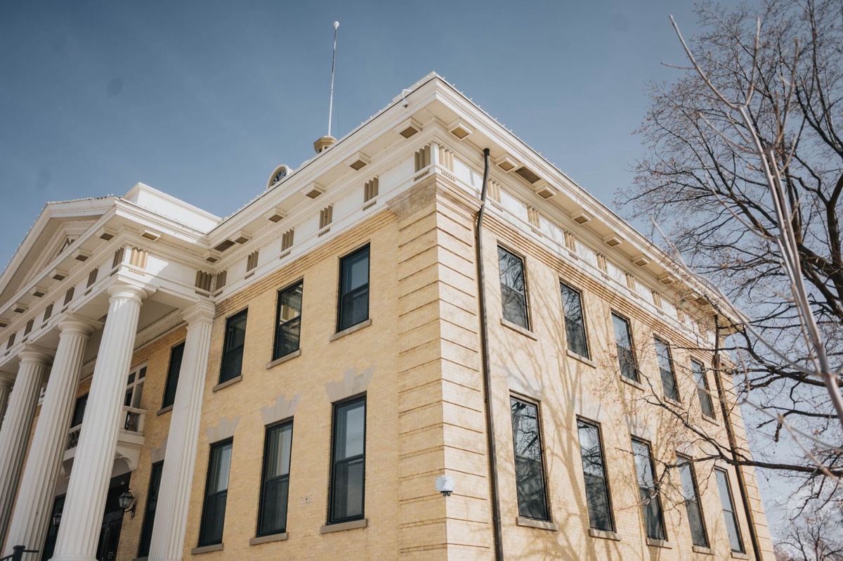 Low-angle view of the corner of a classical yellow-brick courthouse with white columns and cornice against a clear blue sky, framed by bare tree branches.