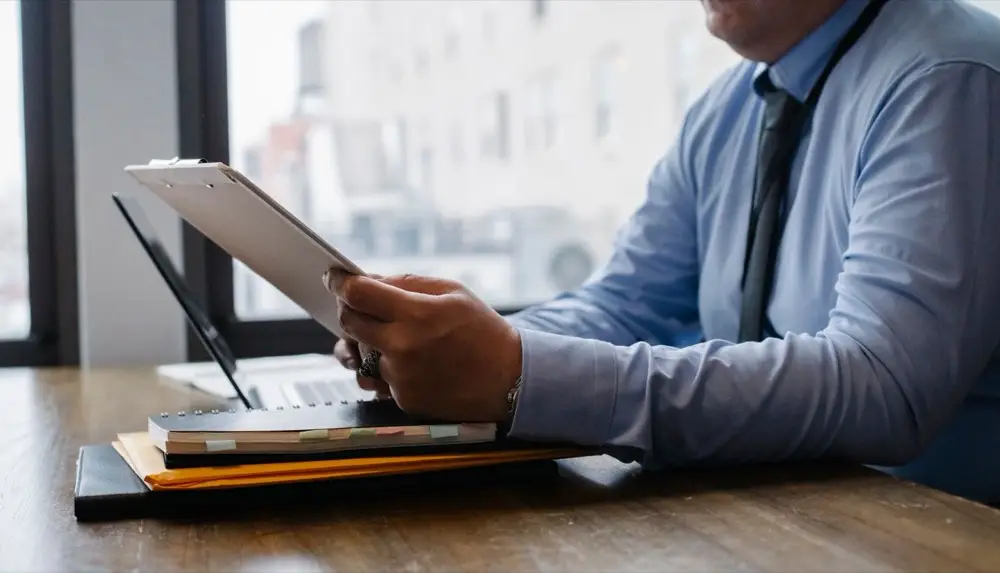 A man in a blue shirt and black tie sitting at a wooden desk in an office, reading paperwork attached to a clipboard.