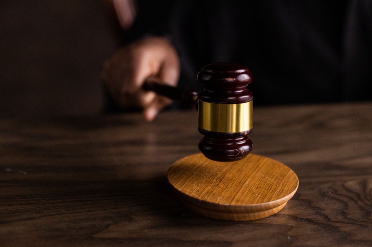 A dark wooden gavel with a gold band, held mid-strike above its round sound block on a dark wooden table.