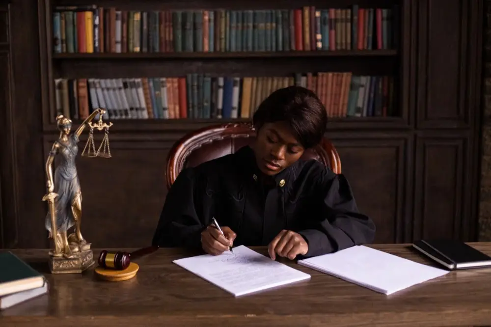 A judge in black robes seated at a desk in a law library, writing on a document surrounded by law books.