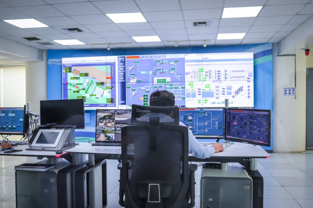 An operator seated in a high-backed chair in a brightly lit control room, working across several desktop monitors in front of a wall-sized display of system dashboards.