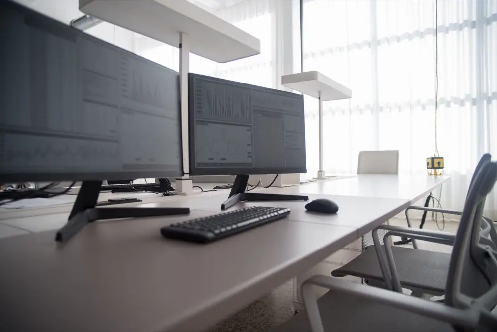 A tidy contemporary office workspace with two large monitors, a keyboard, and ergonomic chairs by a window.