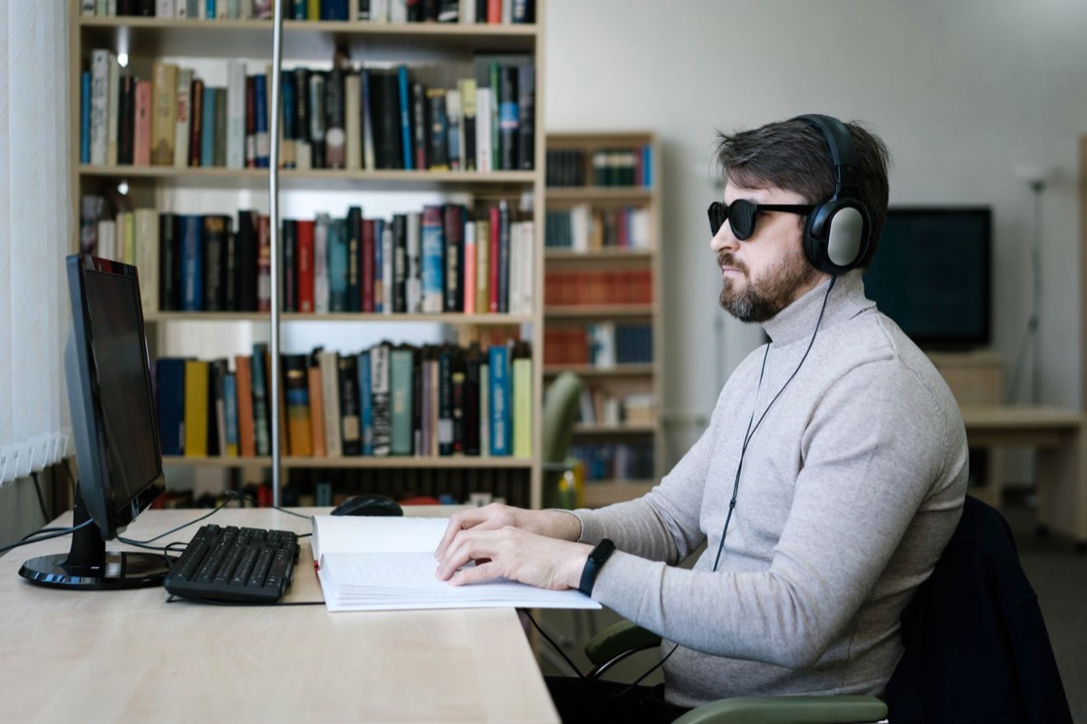A man wearing dark glasses and over-ear headphones sits at a library desk, reading a braille document with his fingers next to a computer monitor and keyboard.