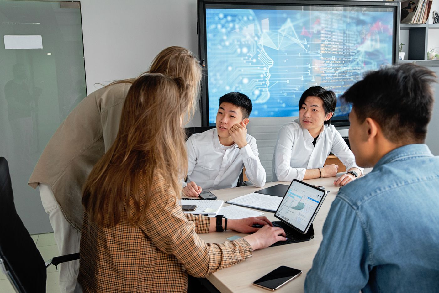 A team of five professionals in a conference room meeting around a table with laptops and papers, in front of a large wall screen displaying code and data visualizations.