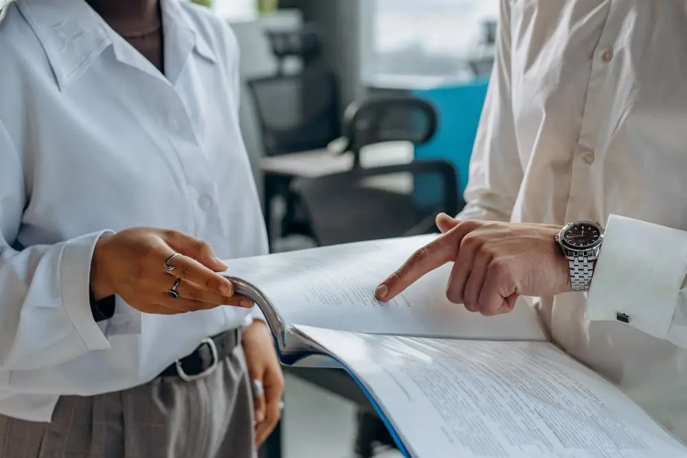Close-up of two colleagues in white shirts standing together, one pointing at a line of text in an open binder of printed report pages.