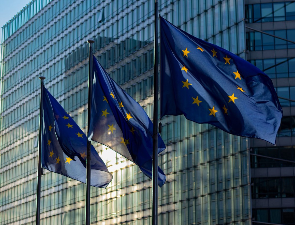 Three European Union flags waving on poles in front of the glass facade of the European Commission building in Brussels.