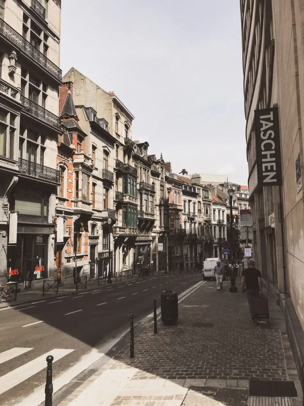 A quaint Brussels street lined with historic brown-brick buildings, with pedestrians on the sidewalk and cars parked along the road under a bright sky.