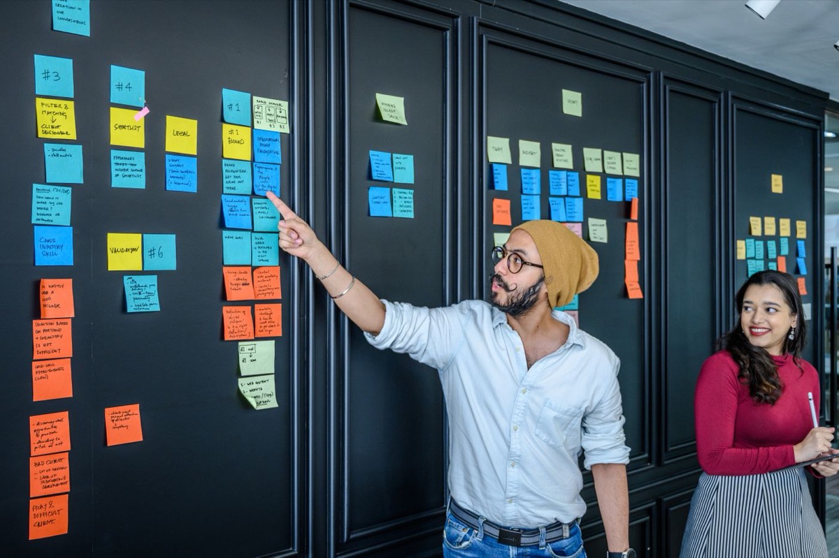 A man in a beanie points at one of dozens of colorful handwritten sticky notes arranged on a dark wall, while a smiling colleague with a notepad looks on.