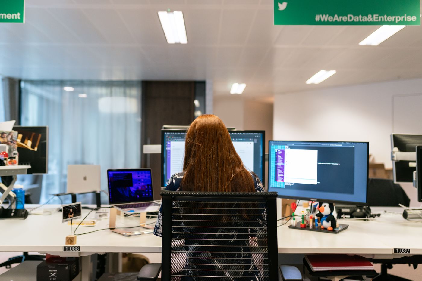 A woman with long red hair seen from behind at an office desk, working across two large monitors and a laptop displaying code and developer tools.