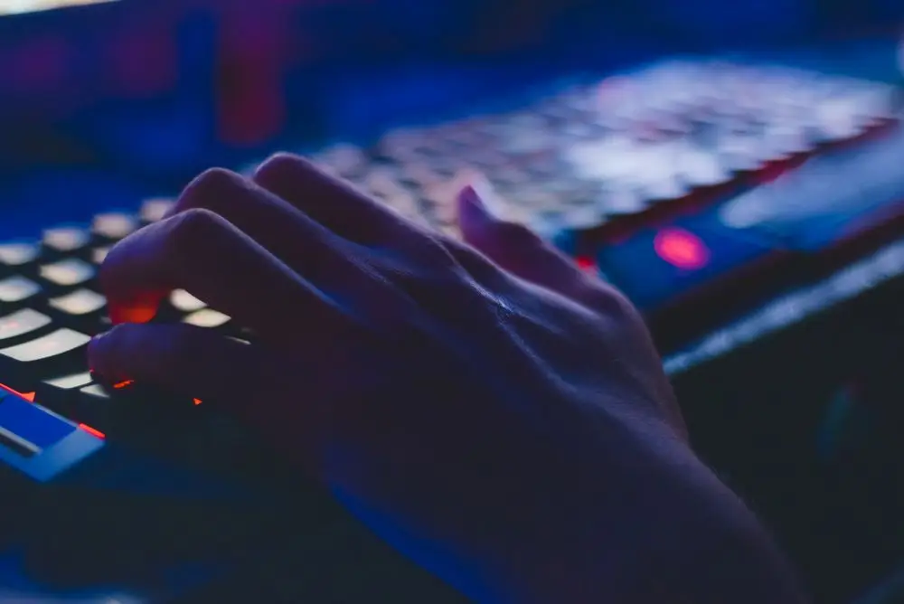 Close-up of a hand resting on a backlit mechanical keyboard in a dark room, lit by the blue glow of a computer screen.
