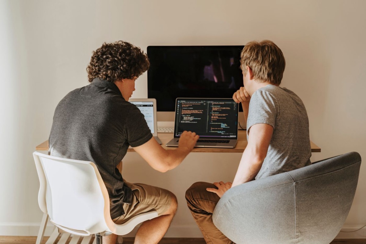 Two young developers sitting side by side at a small desk, leaning in and reviewing code together on a laptop screen.
