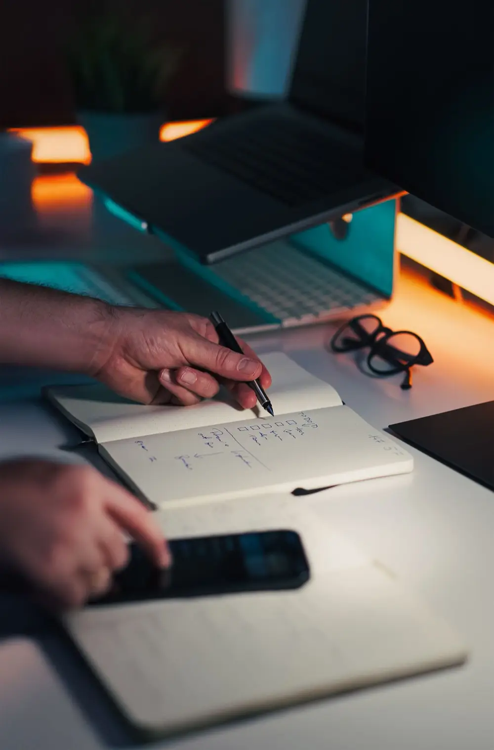 Close-up of a person's hands writing notes in a notebook on a sleek desk, with a laptop and smartphone nearby.
