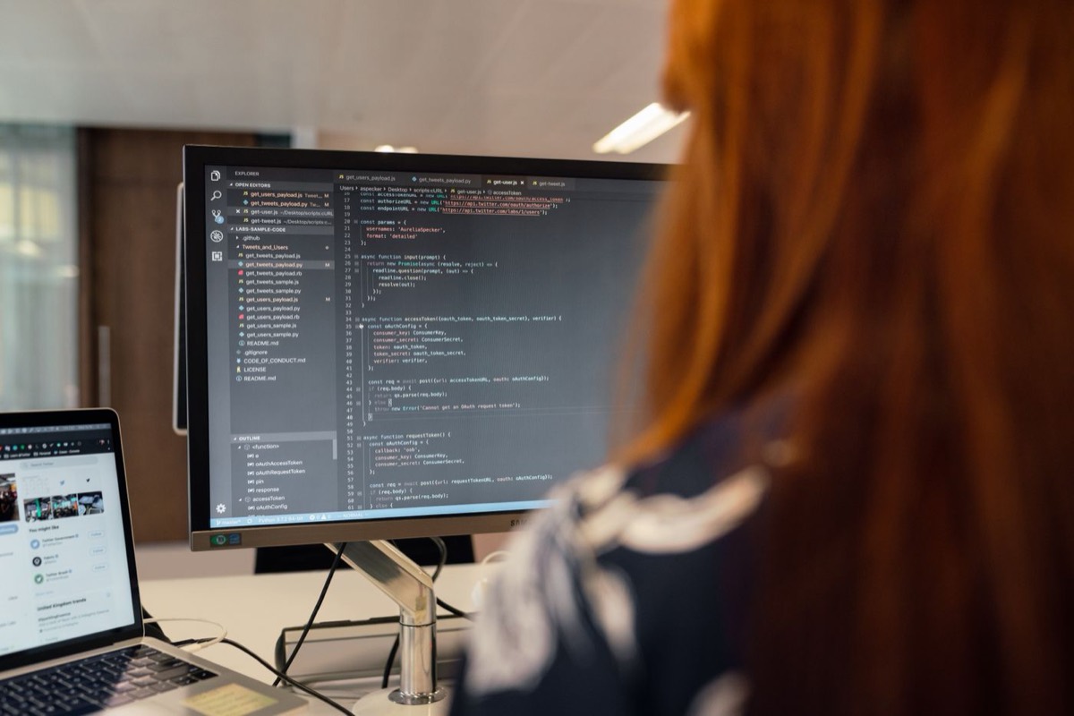 A developer with long red hair seen from behind, working at a desk in a bright office with a code editor full of JavaScript open on a large monitor and a laptop beside it.