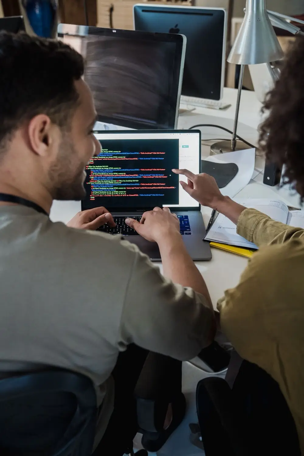 A man and a woman sitting together at a laptop in a modern office, talking through the code displayed on the screen.
