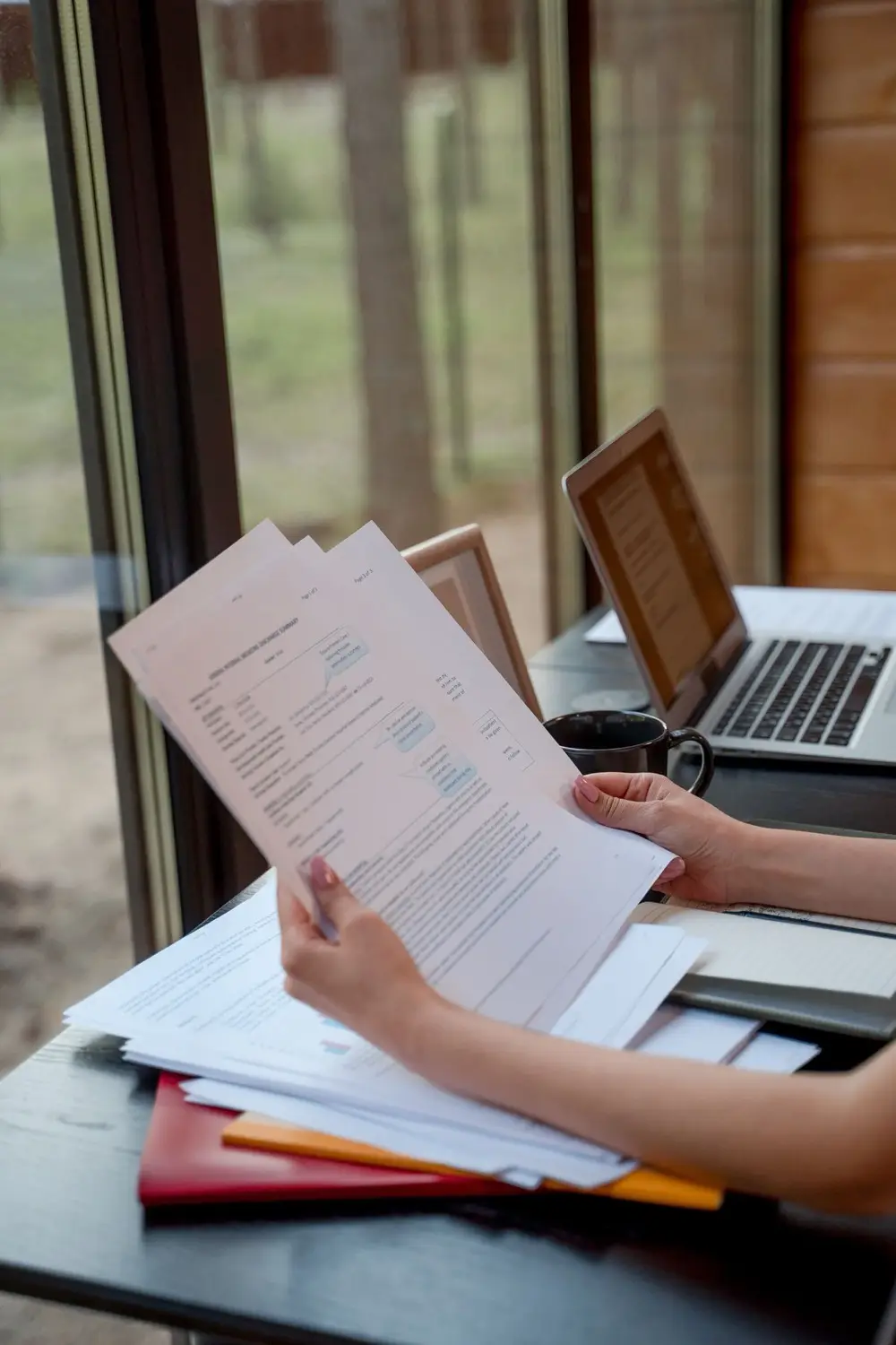 A person standing near a window holding and reviewing printed documents, with open laptops on the desk in front of them.