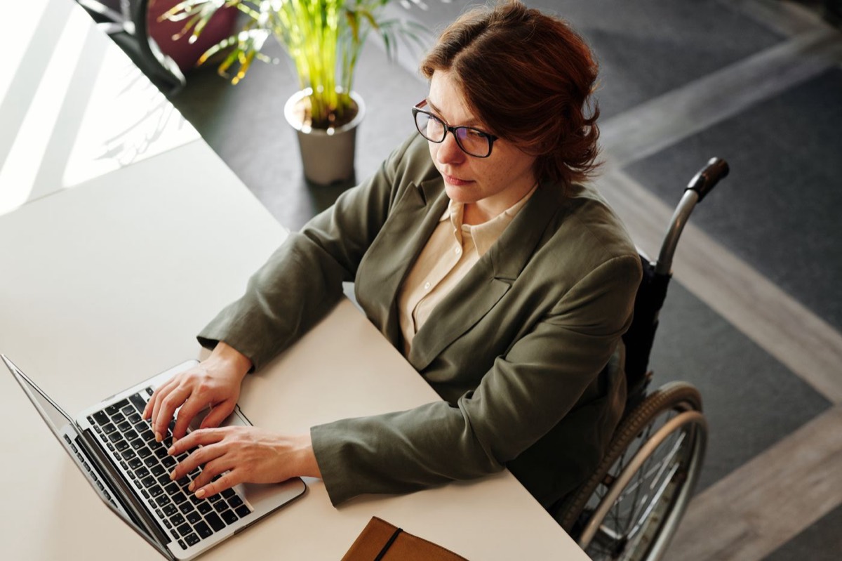 Seen from above, a woman with glasses seated in a wheelchair types on a laptop at a white office desk with a potted plant beside her.