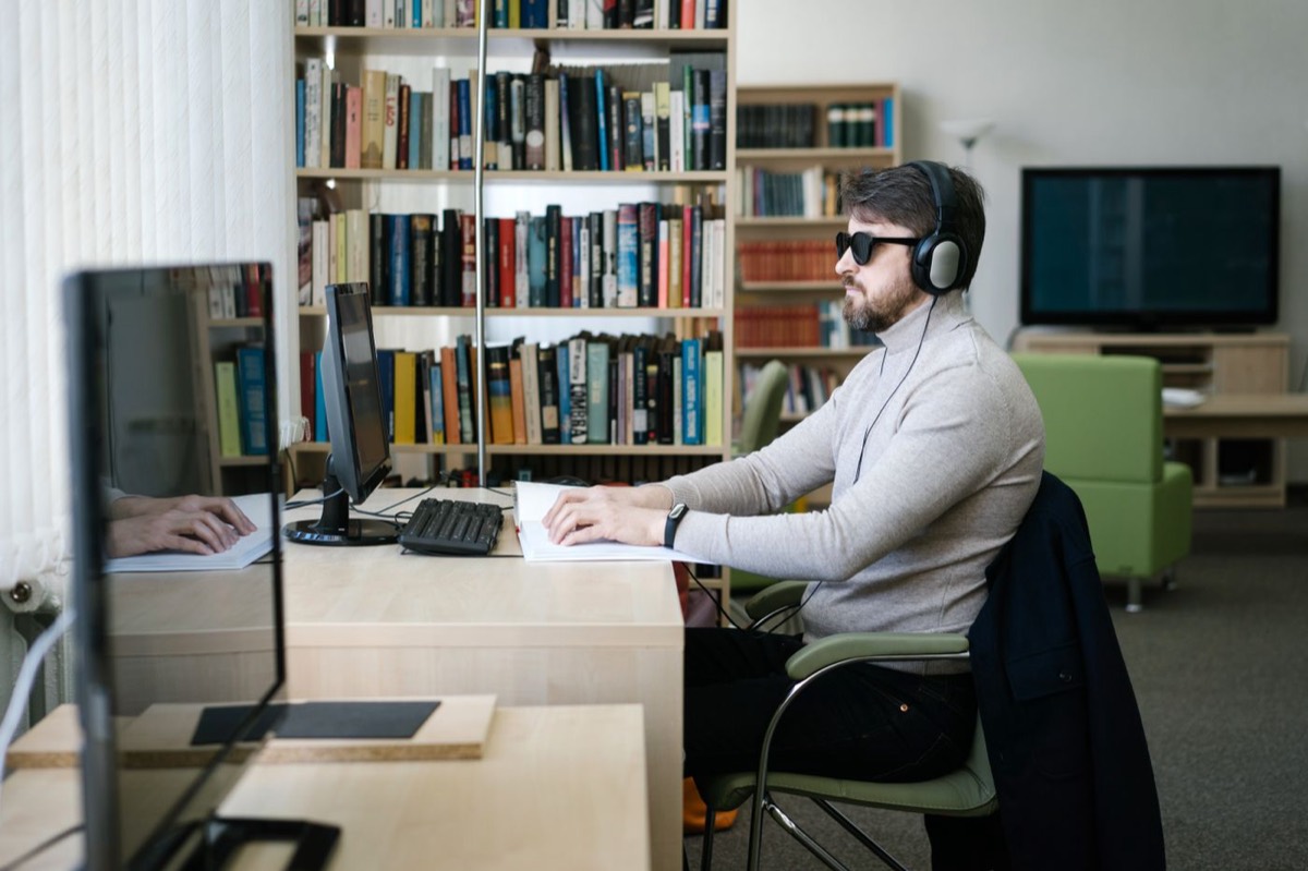A man wearing dark glasses and headphones works at a library computer, one hand on a braille document beside the keyboard and monitor.