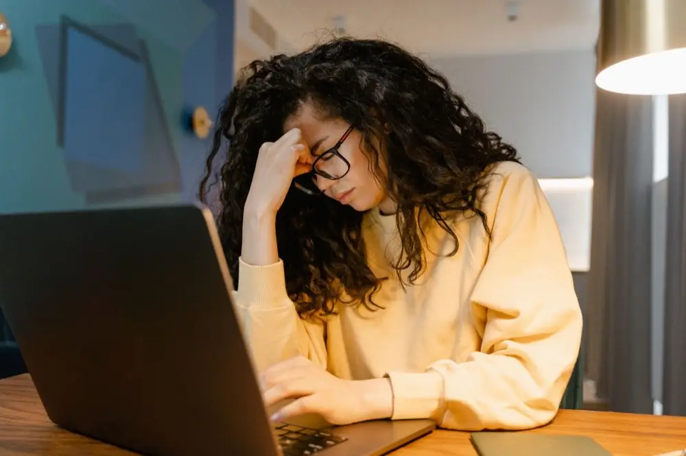 A woman with curly hair and glasses looks strained and tired while working on a laptop.